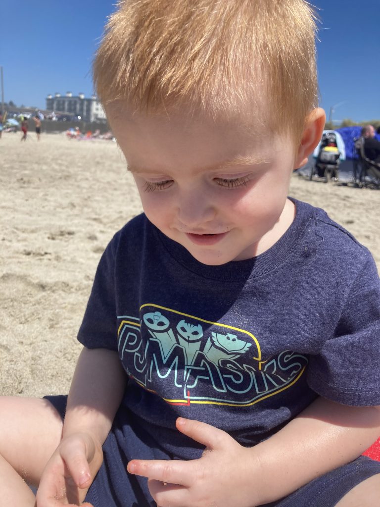 A 3-year-old little boy sitting on the beach. He is wearing a blue shirt that says PJ Masks and smiling at his sandy hands.